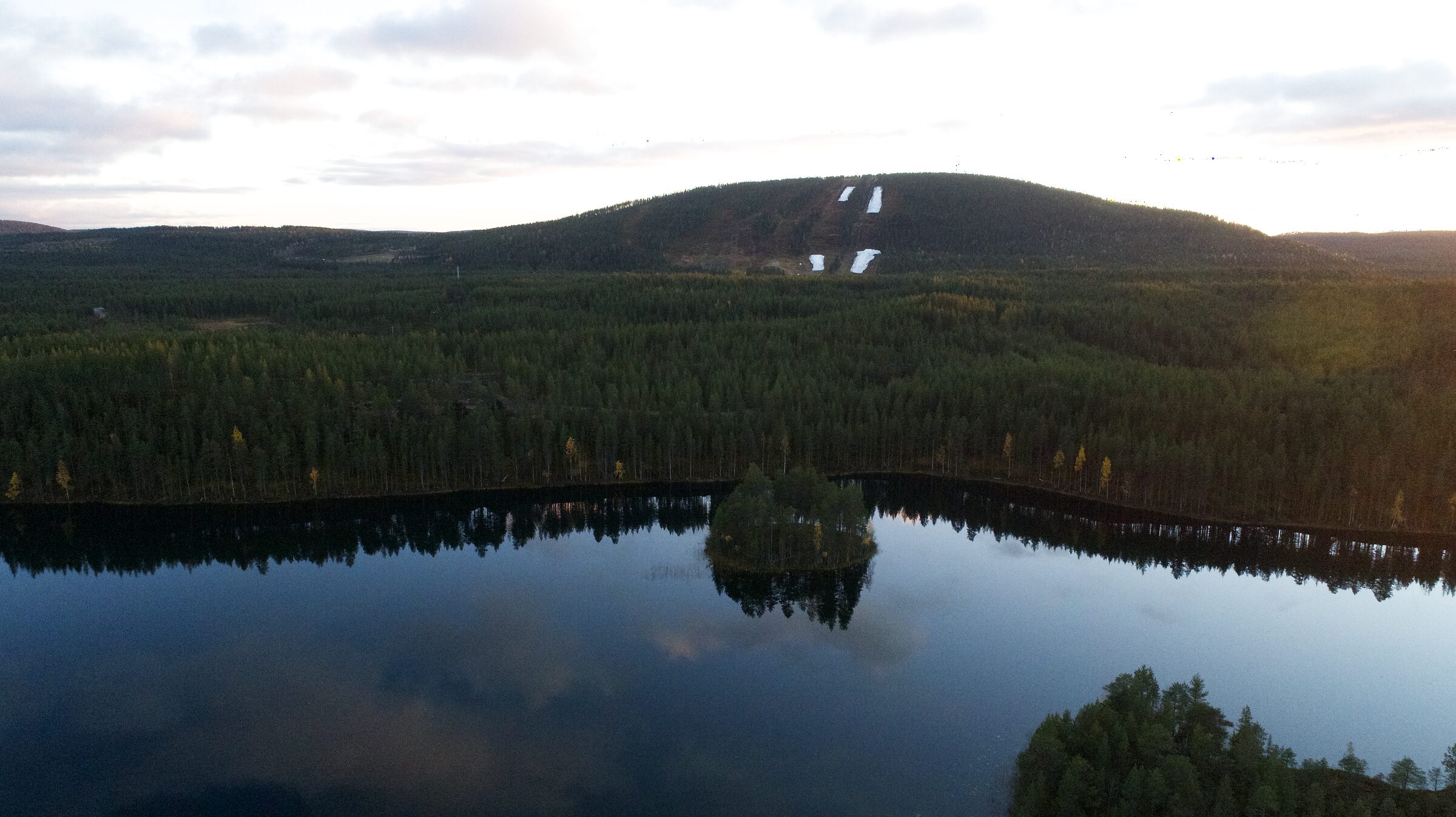 Ilmakuva Herajärvestä ja sen saaresta, takana mäntymetsää ja Suomutunturi rinteineen. Hera Resortin sijainti on merkitty järven takarannalle puiden keskelle.